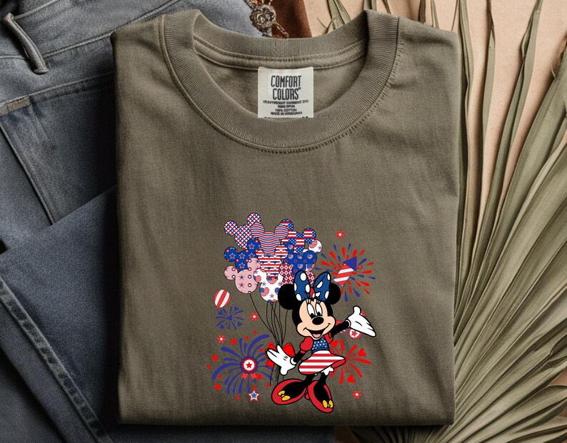K&ouml;nnte beinhalten: Ein khakifarbenes T-Shirt mit einer Grafik einer Zeichentrickmaus in einem patriotischen Kleid, die ein B&uuml;ndel Luftballons in Form von Mausohren mit rot-wei&szlig;-blauen Mustern h&auml;lt, umgeben von Illustrationen von Feuerwerksk&ouml;rpern.