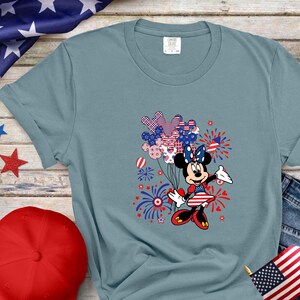 K&ouml;nnte beinhalten: Ein schieferblaues Kurzarm-T-Shirt mit einer Grafik von Minnie Maus, die ein B&uuml;ndel Luftballons mit rot-wei&szlig;-blauem Sternen- und Streifenmuster h&auml;lt, umgeben von Feuerwerksdarstellungen in den passenden patriotischen Farben.