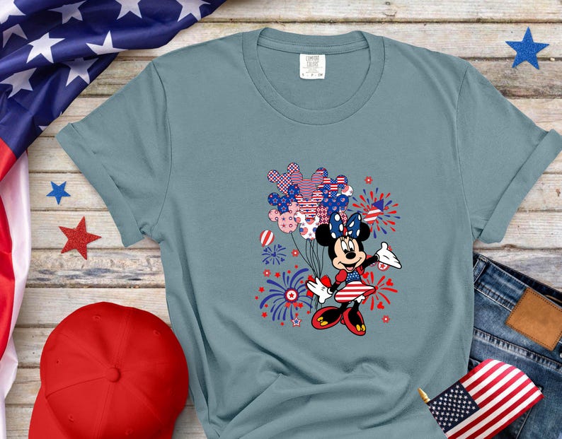 K&ouml;nnte beinhalten: Ein schieferblaues Kurzarm-T-Shirt mit einer Grafik von Minnie Maus, die ein B&uuml;ndel Luftballons mit rot-wei&szlig;-blauem Sternen- und Streifenmuster h&auml;lt, umgeben von Feuerwerksdarstellungen in den passenden patriotischen Farben.