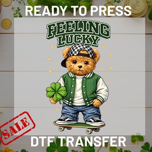 Peut inclure: Transfert DTF avec un ours en peluche portant une veste verte et une casquette à carreaux, tenant un trèfle à quatre feuilles. L'ours est sur un skateboard, avec le texte "FEELING LUCKY" et "DTF TRANSFER".