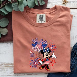 K&ouml;nnte beinhalten: Ein terrakottafarbenes T-Shirt mit einem Comfort Colors-Etikett am Halsausschnitt. Die Grafik zeigt eine Zeichentrickmaus in einem patriotischen Kleid mit Schleife, umgeben von Illustrationen roter, wei&szlig;er und blauer Luftballons und Feuerwerke.