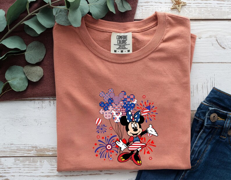 K&ouml;nnte beinhalten: Ein terrakottafarbenes T-Shirt mit einem Comfort Colors-Etikett am Halsausschnitt. Die Grafik zeigt eine Zeichentrickmaus in einem patriotischen Kleid mit Schleife, umgeben von Illustrationen roter, wei&szlig;er und blauer Luftballons und Feuerwerke.