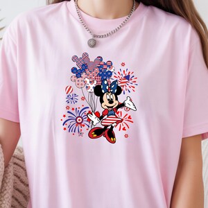 K&ouml;nnte beinhalten: Ein hellrosa T-Shirt mit einem Grafikdruck einer Zeichentrickmaus, die ein patriotisches Kleid mit roten, wei&szlig;en und blauen Sternen und Streifen tr&auml;gt. Das Design zeigt zudem eine Ansammlung von Luftballons und Feuerwerk.