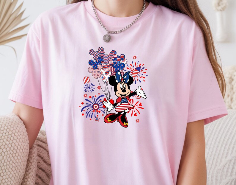 K&ouml;nnte beinhalten: Ein hellrosa T-Shirt mit einem Grafikdruck einer Zeichentrickmaus, die ein patriotisches Kleid mit roten, wei&szlig;en und blauen Sternen und Streifen tr&auml;gt. Das Design zeigt zudem eine Ansammlung von Luftballons und Feuerwerk.