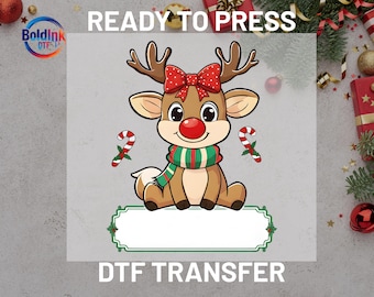 May include: A cartoon reindeer with a red nose, a red bow, and a green and red scarf. Two candy canes flank the reindeer. The text "READY TO PRESS" and "DTF TRANSFER" are at the top and bottom of the image, respectively. A blank label is at the bottom.