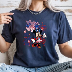 K&ouml;nnte beinhalten: Ein kurz&auml;rmeliges T-Shirt in Marineblau mit einer Grafik von Minnie Maus, die ein B&uuml;ndel Luftballons in Form von Mickey-Maus-K&ouml;pfen h&auml;lt. Das Design enth&auml;lt rote, wei&szlig;e und blaue Farben mit Sternen- und Feuerwerksmustern.