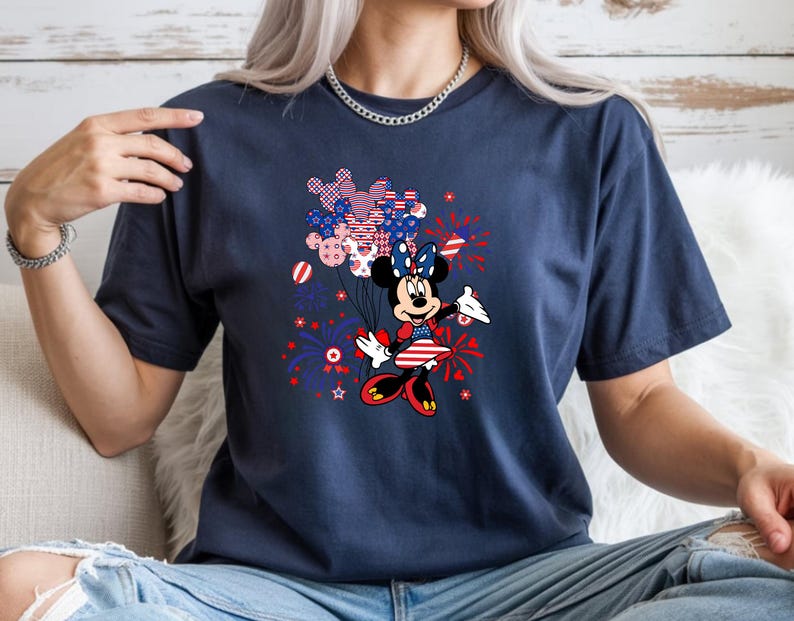 K&ouml;nnte beinhalten: Ein kurz&auml;rmeliges T-Shirt in Marineblau mit einer Grafik von Minnie Maus, die ein B&uuml;ndel Luftballons in Form von Mickey-Maus-K&ouml;pfen h&auml;lt. Das Design enth&auml;lt rote, wei&szlig;e und blaue Farben mit Sternen- und Feuerwerksmustern.