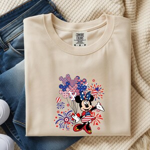 K&ouml;nnte beinhalten: Ein beiges T-Shirt mit Rundhalsausschnitt und Comfort Colors-Etikett, das eine Comic-Mausfigur in einem patriotischen Kleid mit Schleife zeigt, die ein B&uuml;ndel Luftballons mit Sternen- und Streifenmuster h&auml;lt, umgeben von roten und blauen Feuerwerken.