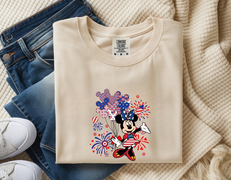 K&ouml;nnte beinhalten: Ein beiges T-Shirt mit Rundhalsausschnitt und Comfort Colors-Etikett, das eine Comic-Mausfigur in einem patriotischen Kleid mit Schleife zeigt, die ein B&uuml;ndel Luftballons mit Sternen- und Streifenmuster h&auml;lt, umgeben von roten und blauen Feuerwerken.