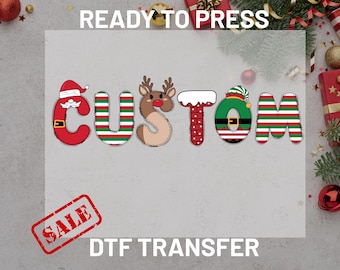 May include: A festive DTF transfer design with the word "CUSTOM" spelled out in Christmas-themed letters. The letters are red, green, and brown, with Santa, reindeer, and elf hat designs. The word "READY TO PRESS" is at the top, and "SALE" and "DTF TRANSFER" are at the bottom.