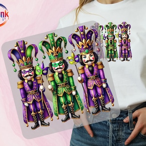 May include: White t-shirt featuring a colorful graphic of three nutcracker figures in purple and green jester costumes, holding masks and staffs. The figures have elaborate hats and are set against a light background. The BoldInk QTF logo is in the upper left corner.