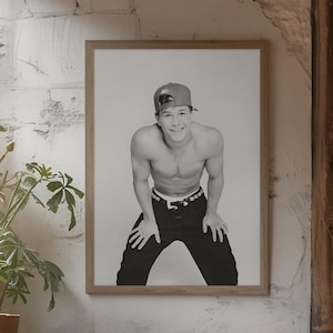 May include: Black and white framed print of a person wearing a baseball cap and black pants. The person is smiling and leaning forward. The print is hanging on a white textured wall. A small table and chairs are in front of the print.