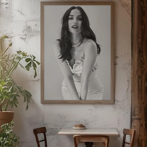 May include: A framed black and white photograph of a woman in a light-colored dress and necklace. The photograph is hanging on a textured white wall. The scene includes a small table, two chairs, and a potted plant.