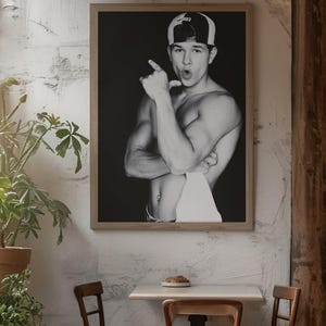 May include: Black and white framed print of a person wearing a baseball cap, pointing with one hand and with the other hand crossed over their chest. The print is hanging on a white textured wall. The image is in a cafe setting with a table and chairs.