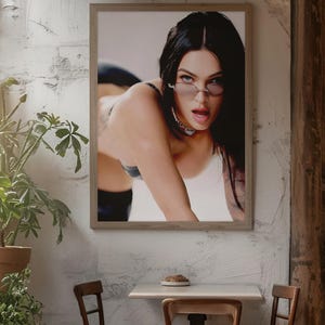 Puede incluir: Una impresión enmarcada que presenta a una persona con gafas de sol y un collar de gargantilla en una pose sugerente. La impresión se muestra en una pared blanca texturizada, con una pequeña mesa y sillas al frente.