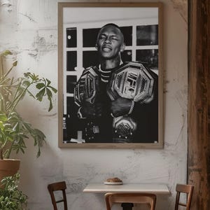 May include: Black and white framed print of a man holding two championship belts. The man is wearing a necklace and has a serious expression. The belts have the UFC logo. The print is displayed in a cafe setting.
