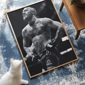 May include: Black and white framed print featuring a shirtless athlete with tattoos, wearing UFC gloves and shorts. The print is on a blue and white patterned rug, with a white cat in the foreground.