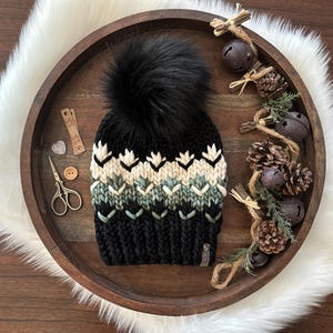 May include: A black knit beanie with a large black faux fur pom-pom. The hat features a geometric pattern in white, beige, and green. The hat is displayed on a wooden tray with decorative elements, including bells and pinecones.