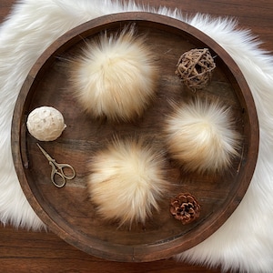 May include: Three fluffy faux fur pom poms in various shades of cream and beige displayed on a wooden tray with decorative accents.