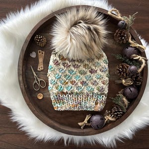 May include: A cream-colored knit hat with a large, fluffy, light brown faux fur pom-pom. The hat features a textured pattern with teal, purple, and brown accents. The hat is displayed on a wooden tray with decorative elements.