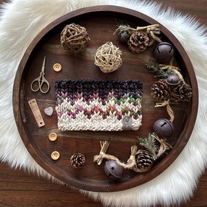 May include: A hand-knitted, multi-colored cowl with a gradient design, featuring shades of black, purple, green, and white. The cowl is displayed on a wooden tray with decorative elements like pinecones, bells, and buttons. The tray sits on a white faux fur surface.