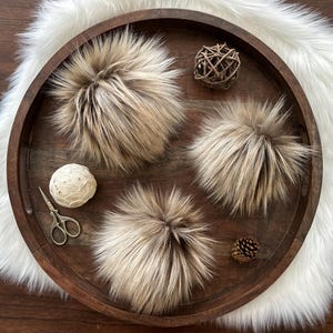 May include: Three fluffy faux fur pom poms in shades of brown and beige, displayed on a wooden tray with a white faux fur background.