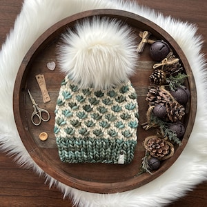 May include: A hand-knitted winter hat with a cream-colored pom-pom and a teal and cream geometric pattern. The hat sits in a wooden tray with decorative elements like pinecones, bells, and faux fur.