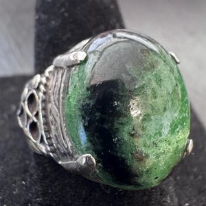 May include: A silver ring with a large, oval-shaped green stone. The stone has dark green and black inclusions. The ring has a decorative silver band with an openwork design on the side.