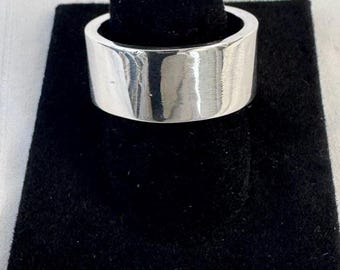 May include: A wide, silver-colored ring with a flat top and sides. The ring is displayed on a black velvet ring holder against a white background. The ring has a smooth, polished surface.