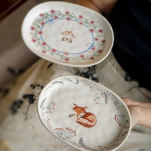 Peut inclure: Deux plats de service ovales en céramique. L'un présente une bordure florale avec un ruban bleu et une illustration de chat. L'autre présente une illustration de renard endormi entourée de motifs botaniques. Les deux sont peints à la main.