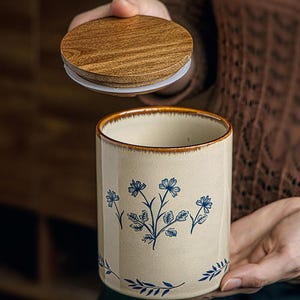 May include: A beige ceramic jar with a wooden lid. The jar features blue floral designs and a brown rim. The lid is round and made of wood. The jar is cylindrical and appears to be a container for kitchen or home use.