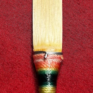 Puede incluir: Primer plano de una caña para instrumento de viento. La caña tiene una punta rectangular de color claro y una base colorida envuelta con hilos rojos, verdes y amarillos. El fondo es una tela roja lisa.