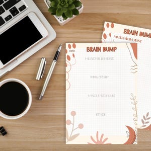 Pode incluir: Uma cena de escritório com um laptop, smartphone, caneta e xícara de café. Dois blocos de notas "Brain Dump" são visíveis, com seções para casa, trabalho e autocuidado. Os blocos têm um padrão de grade e elementos florais.