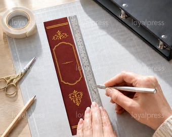 May include: A rectangular dark red bookmark featuring gold filigree accents and a central blank frame, positioned next to a metal ruler, a roll of white tape, a pencil, and gold scissors on a gray cutting mat with a black binder nearby.