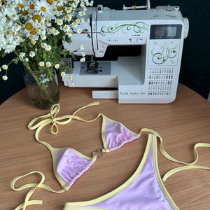 May include: A two-piece bikini set featuring light purple fabric with pale yellow trim. The top has triangle cups connected by a silver heart-shaped ring, and the bottom is a high-cut style with side ties. Both pieces include matching yellow straps.