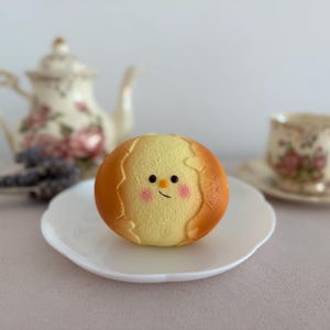 May include: A round, yellow, textured bread roll with a brown crust edge, featuring a printed cartoon face with black eyes, a small orange nose, and pink cheeks, centered on a white scalloped plate with a floral teapot and teacup nearby.