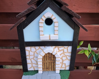 May include: A decorative birdhouse with a light blue facade, black trim, and a brown shingled roof. The birdhouse features a stone-like lower section with a wooden door and a gray stone-like platform extending outward.