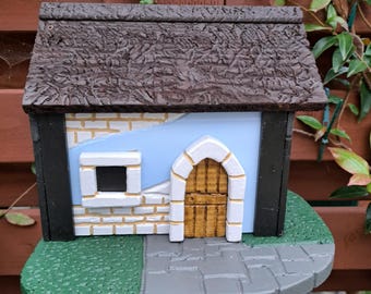 May include: A decorative birdhouse with a brown textured roof, white brick walls, and a light blue background. The birdhouse features a small window and a wooden arched door. The base is green with a gray stone path.