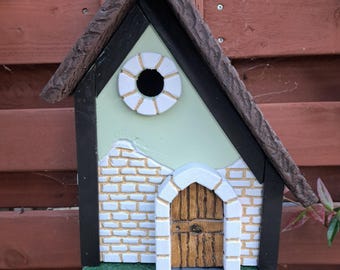 May include: A decorative birdhouse with a church-like design. The birdhouse features a light green roof, a white and gold circular window, and a brick facade with a wooden door. The roof is brown and textured, and the base has a green lawn and gray stone path.