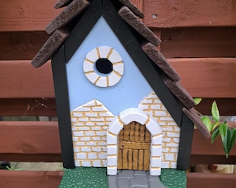 May include: A decorative birdhouse with a whimsical design. The birdhouse features a blue sky background, a white arched doorway, and a brown shingled roof. The facade is painted with white brick and a gray stone path.