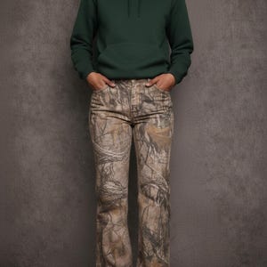 May include: A person wearing a dark green hooded sweatshirt and camouflage print pants. The pants have a brown, tan, and green pattern. The person is standing in front of a gray background.