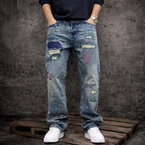 Op de afbeelding: Lichtblauwe denim jeans met distressed details en patches in blauw, paars en wit. De jeans heeft een relaxte pasvorm en wordt gecombineerd met witte sneakers. De stijl is casual en vintage-geïnspireerd.
