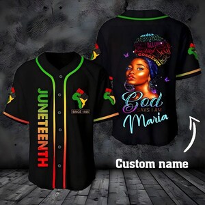 May include: A black baseball jersey with green trim, featuring the word JUNETEENTH in a vertical red-to-yellow gradient, a fist icon, and the text SINCE 1865. The back displays a portrait of a person with text and the phrase God says I am Maria.