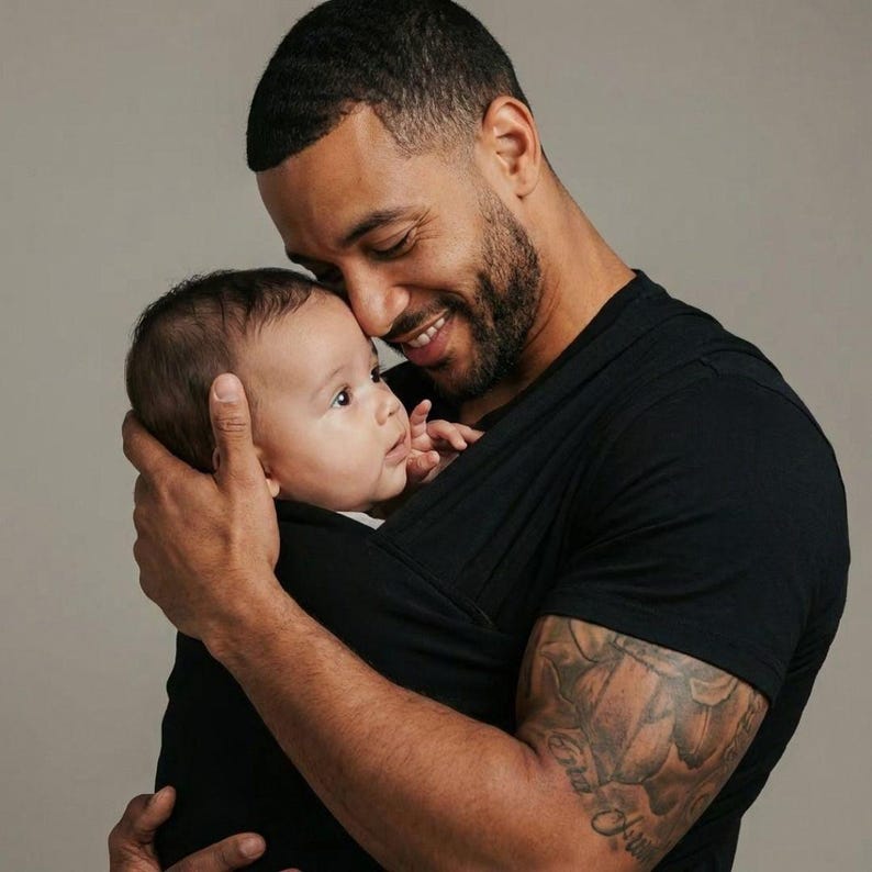 May include: A smiling person holding a baby close. The baby is wrapped in a black carrier. The person is wearing a black t-shirt and has a tattoo on their arm.