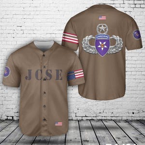 May include: A brown baseball jersey with the text "JCSE" on the front. The jersey features a US flag on the sleeve and a military emblem on the back. The jersey has a button-up front and short sleeves.