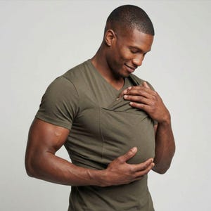 May include: Olive green V-neck shirt with a unique front pocket design. The shirt has short sleeves and a relaxed fit. The fabric appears to be a soft, breathable material, suitable for casual wear. The shirt is designed to accommodate a baby.