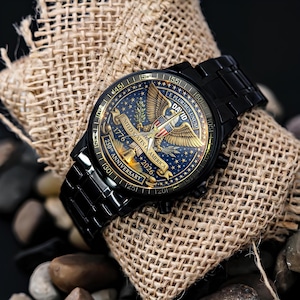 May include: Black wristwatch with a detailed face featuring a gold eagle, stars, and the text "UNITED STATES OF AMERICA 1776-2026 250TH ANNIVERSARY". The watch is set on a burlap sack and surrounded by stones.