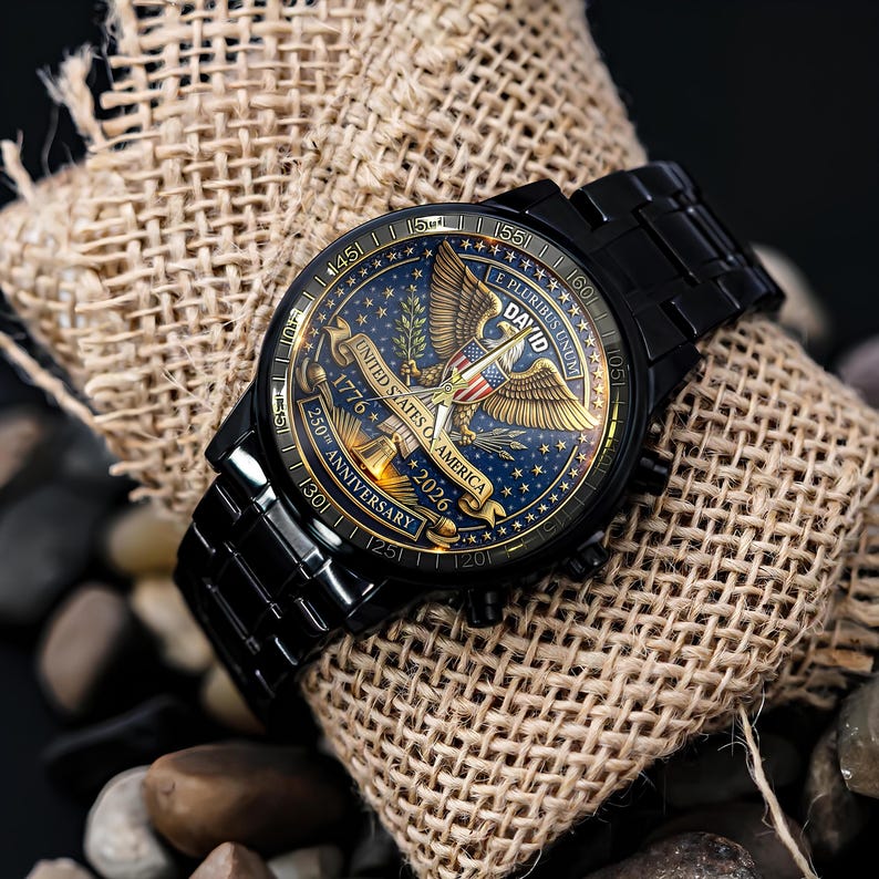 May include: Black wristwatch with a detailed face featuring a gold eagle, stars, and the text "UNITED STATES OF AMERICA 1776-2026 250TH ANNIVERSARY". The watch is set on a burlap sack and surrounded by stones.