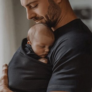 May include: A sleeping baby wrapped in a black fabric carrier, held close by a person wearing a black shirt. The baby's face is visible, with closed eyes and a peaceful expression. The person's face is partially visible, with a beard and a gentle expression.