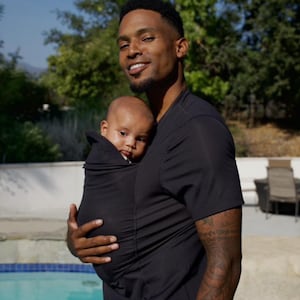 May include: A black baby carrier holds an infant close to a person wearing a black shirt. The baby is wrapped in the carrier, with only the face visible. The background includes greenery and a pool.